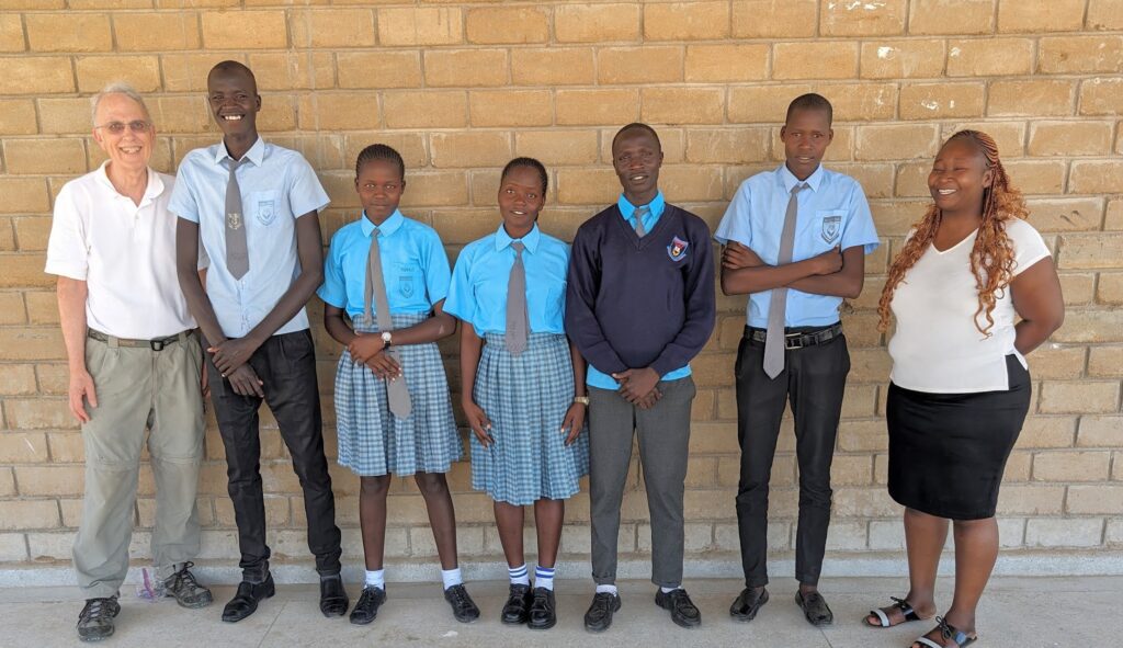 Dale DeJager, Juliet Rabach, and five Joy of Coding students stand with their backs against a brick wall outside of the school. Each student wears a school uniform of a blue button up shirt and gray tie.