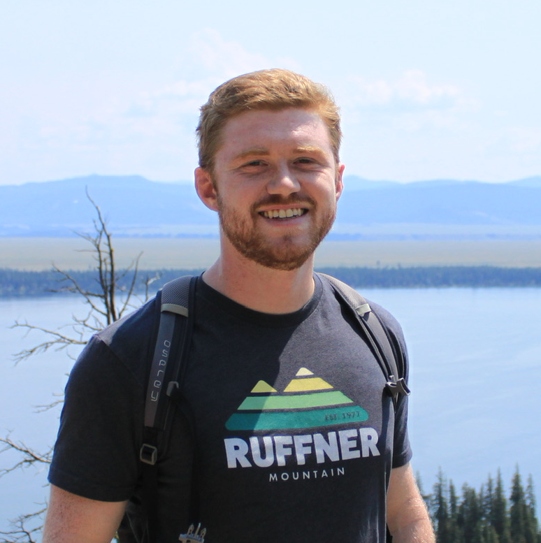 Jacob Sansom wearing a dark tshirt stands smiling at the camera in front of a hilly landscape behind a lake.