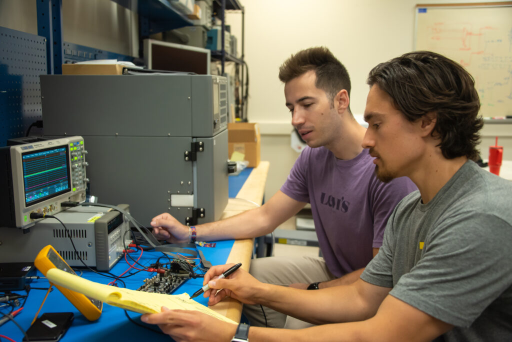 John Kustin shows notes on a legal pad to Vangelis Dikopoulos. They are sitting in their lab, in front of electronic equipment.