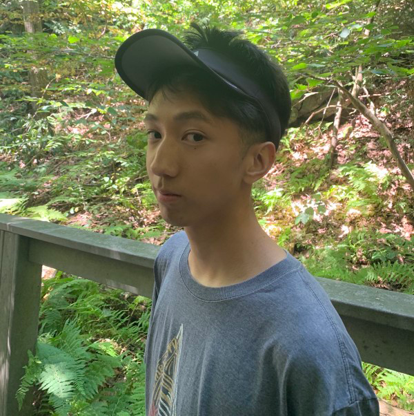 Eric Zhao looks sideways at the camera against a forest background. He is wearing a blue shirt and a black cap.
