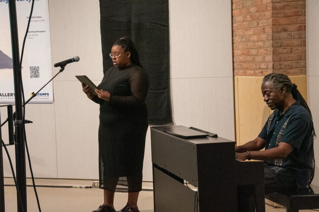 Amber Rogers stands in front of a microphone, singing and reading lyrics from a tablet. Beside her, Herbert Winful plays a piano.