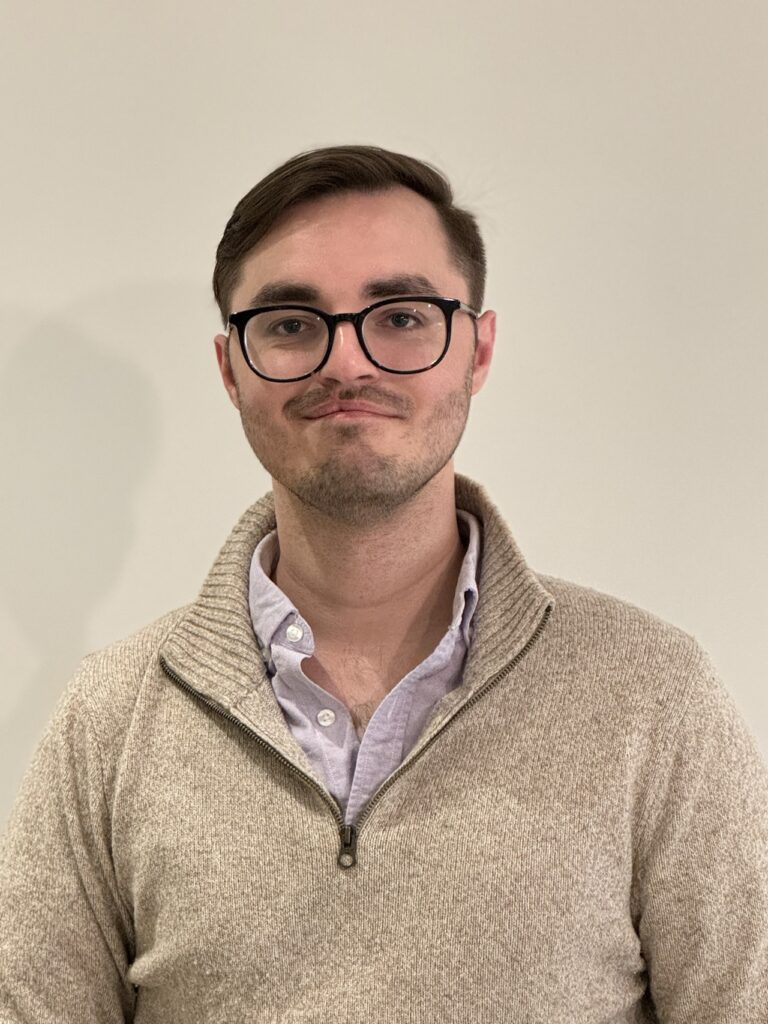 Alexander Bandukwala headshot from mid-chest up. He is wearing a beige quarter-zip sweater with a collared shirt underneath, dark round glasses, and short dark hair.