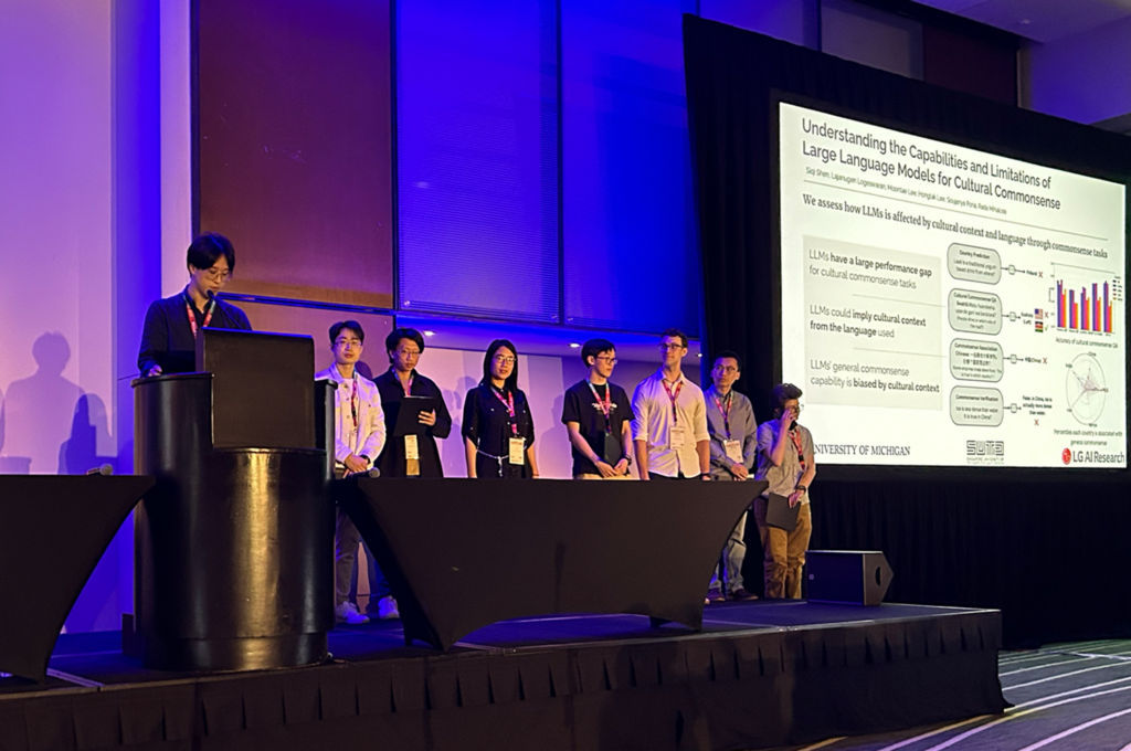 A person speaks at a podium at the front of a dimly lit reception hall. Seven people stand next to him at a table beside the podium. To their left is a slideshow presentation with the first page of the paper described in this article.