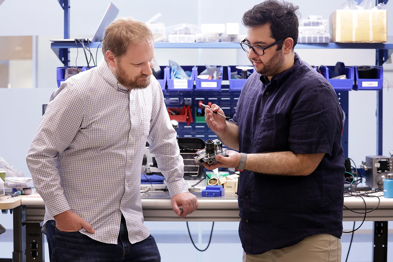 Two men inspect a handheld camera in front of a messy work bench. One man holds a screwdriver and the camera.