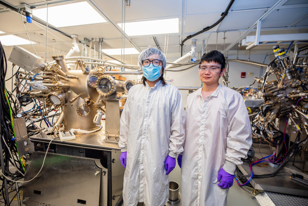 Pan and Zhang stand in a lab. Two devices in the background are full of portholes, pipes and wires.