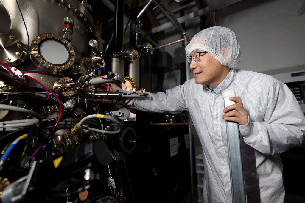 A man wearing a white laboratory coat, eyeglasses, and a white mesh hair cover, stands next to a metal machine with a lot of wires and tubes connected to it.