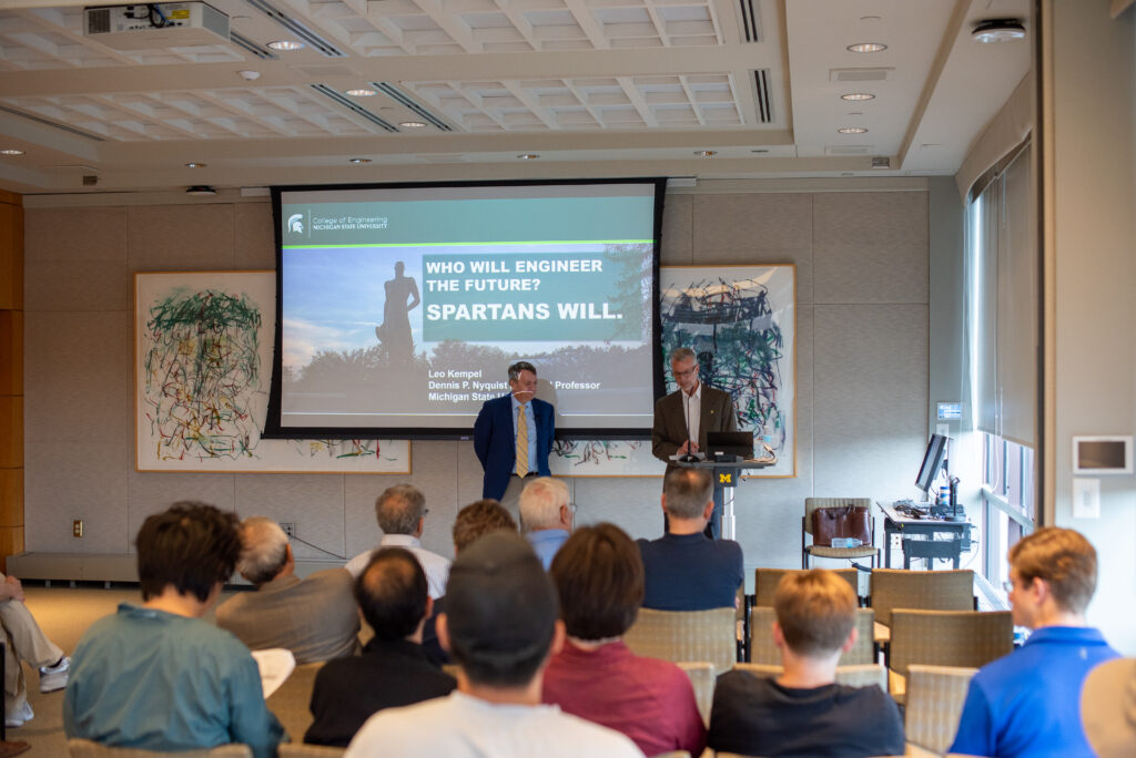 Two men stand at the front of a room - one at a podium and the other in front of a screen with a slide projected that reads: "Who will engineer the future? Spartans will." Rows of chairs filled with audience members are in the foreground of the photo.