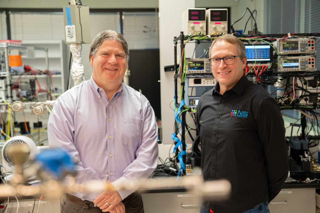 Two men stand side-by-side in a laboratory environment, with electrical equipment behind them. They both wear button down shirts.