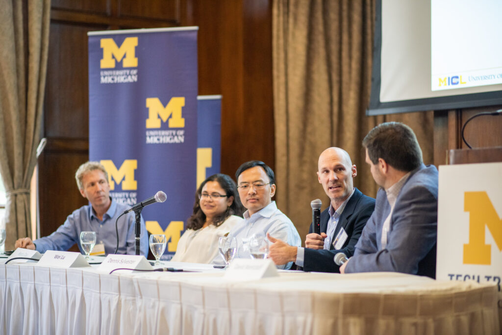 Five panelists sit at a long table on a stage, with University of Michigan signage behind them.