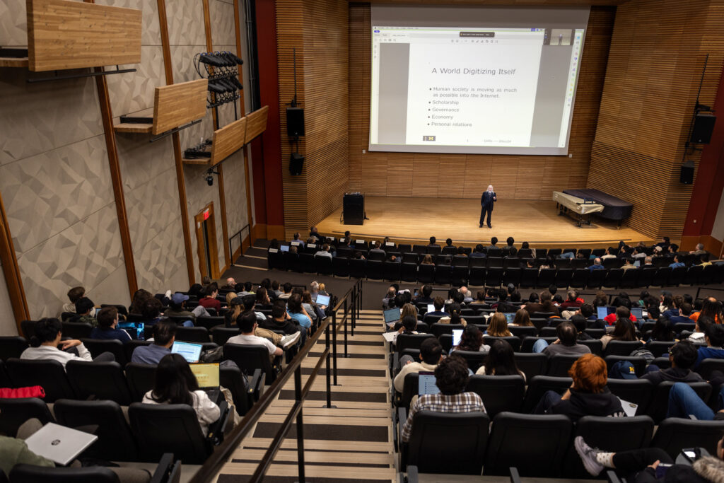 A view of a full auditorium from the back. Walt Diffie stands giving a talk on a wooden stage in the front of the auditorium. Behind him is a large projector screen.