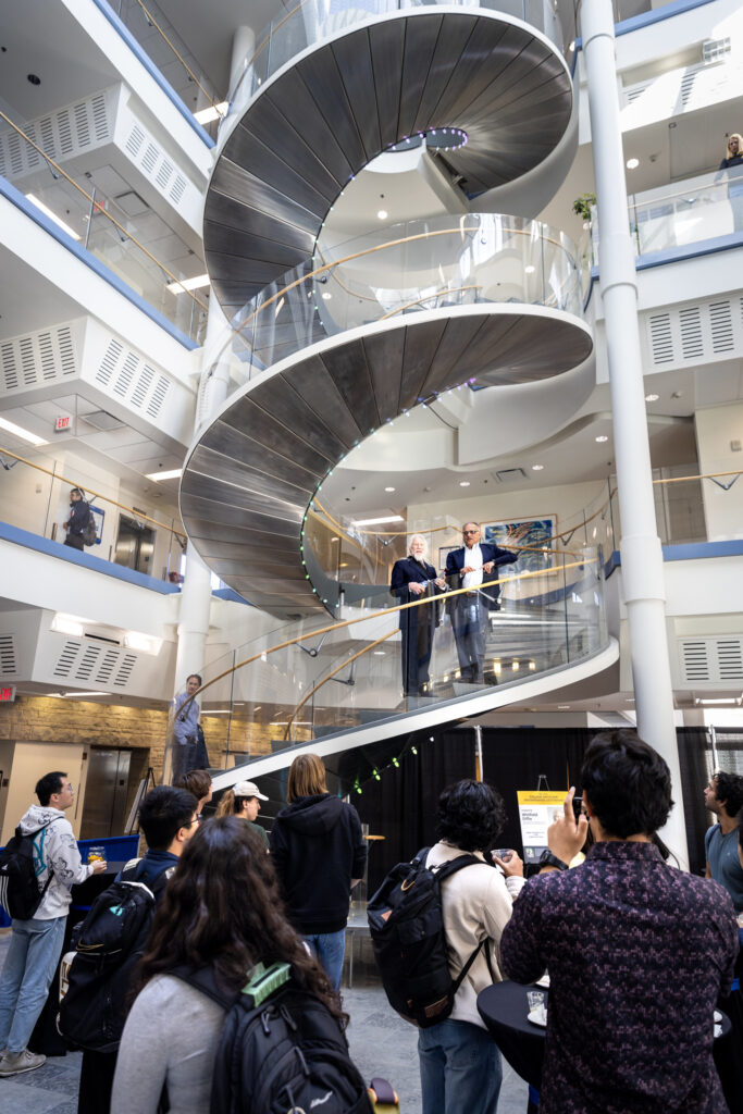 A view of a large spiral staircase from the floor of a light-filled atrium. On the stairs looking down into the atrium stand two figures. Several people stand in the atrium looking up at them, applauding.