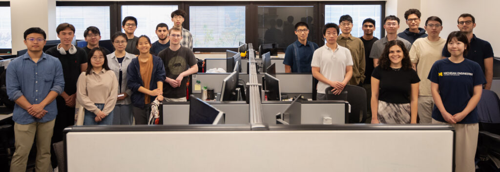 A group photo of 20 students standing in an office room, with a bank of cubicles separating a group of 10 on either side.