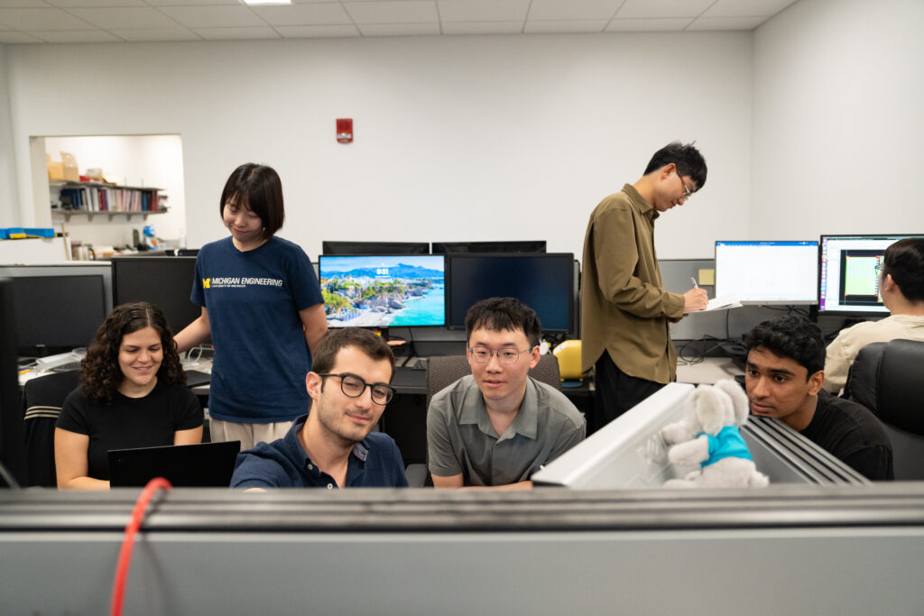 A diverse group of seven students collaborates in an office room, discussing their work, taking notes, and looking at computer screens.