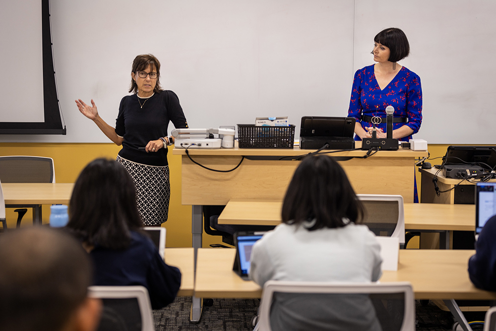 Two women stand at the front of a classroom, talking to a group of students.