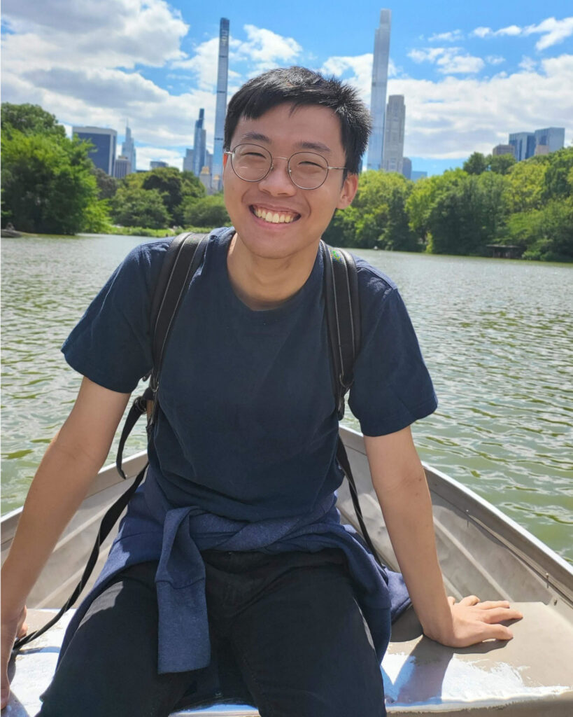 A man sits in a row boat on a body of green-ish water. In the background, a forested park and the skyline of tall skyscrapers is visible.