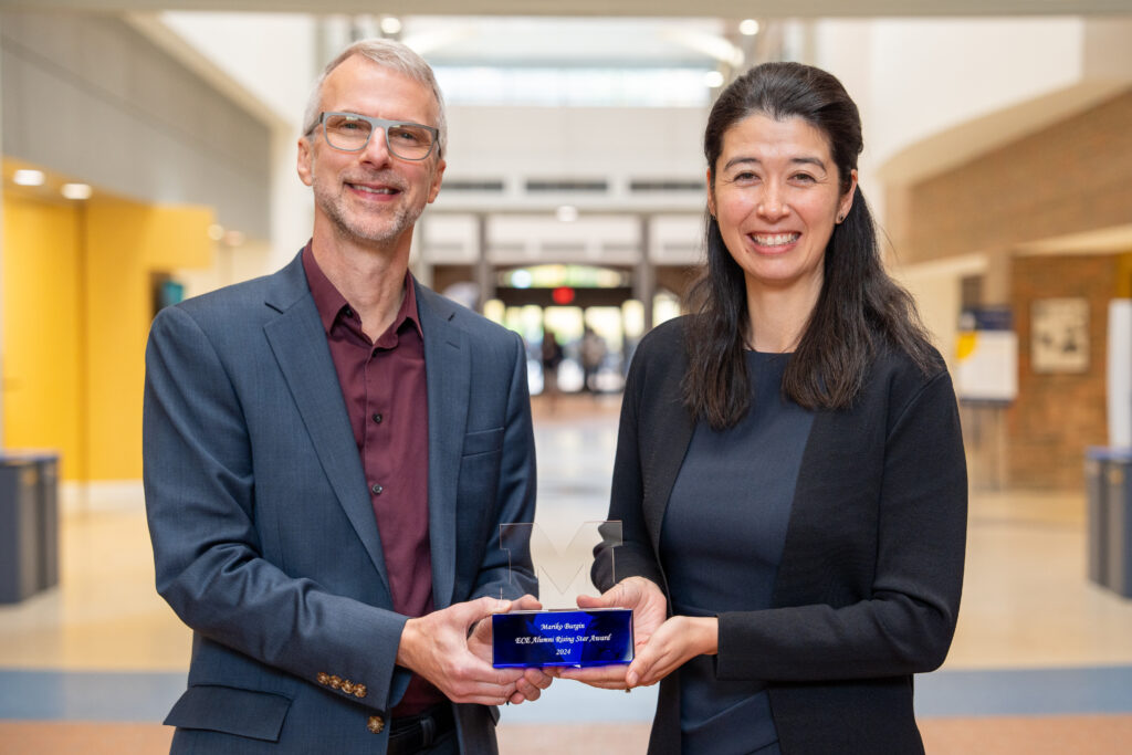 A man and a woman stand side-by-side in the atrium of a building, holding a glass award shaped like the Michigan block M.