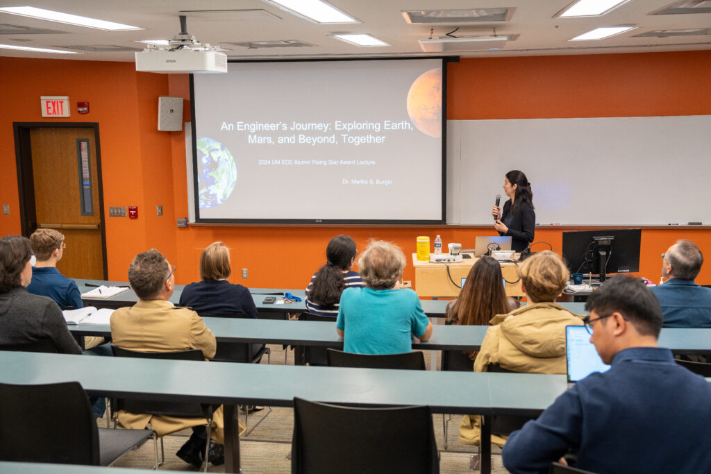 A woman stands in front of an orange classroom, holding a microphone and presenting her lecture to a large audience.