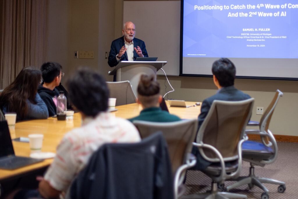 A man stands at the front of a classroom, introducing his talk in front of his title slide.