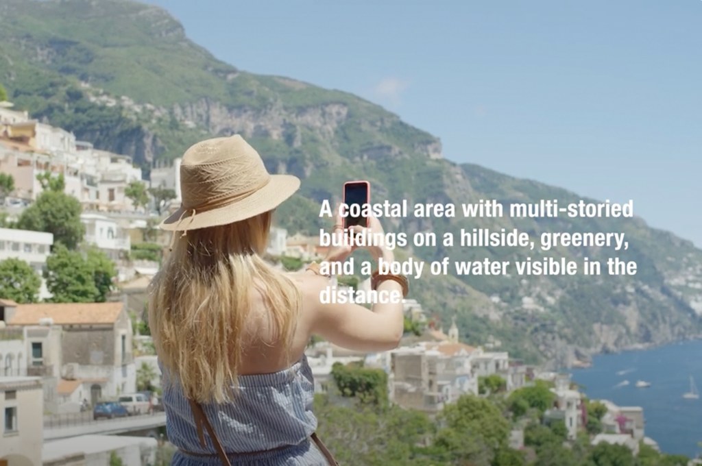 A woman points her phone at a hilly, coastal outlook. A textual description of the scenery overlays the image, reading "A coastal area with multi-storied buildings on a hillside, greenery, and a body of water visible in the distance."