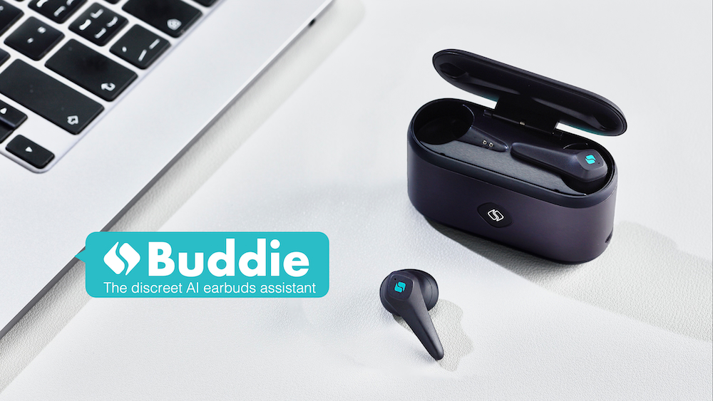 A close up of two black headphones and a black charging case sitting on a desk next to a laptop keyboard. Text overlay reads "Buddie the discreet AI earbuds assistant."