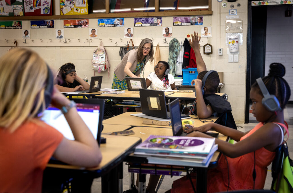 A teacher has walked around an island of four desks to help one of the students sitting at them. The teacher leans over the student's shoulder to point at their laptop screen.