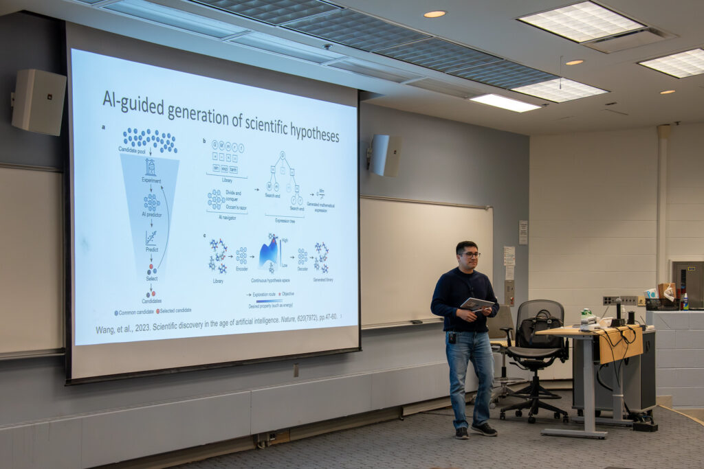 Alex Rodriguez stands at the front of a lecture hall, delivering a lecture. Behind him is a powerpoint slide projected onto a screen with the heading "AI-guided generation of scientific hypotheses"