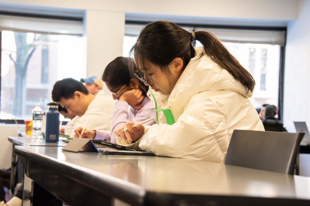 A row of students in a classroom take notes on tablets.