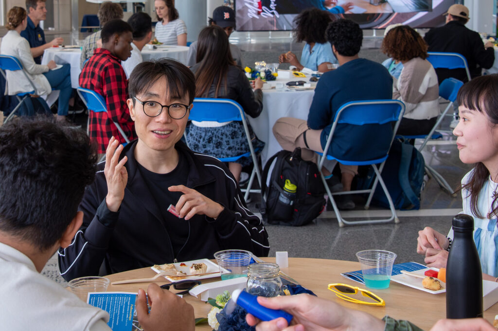 A faculty member seated at a table during an event, talking with students