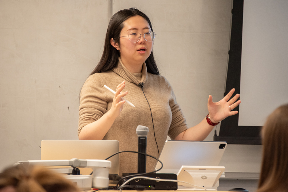 A woman with long black hair and glasses explains a concept to her students, not pictured.