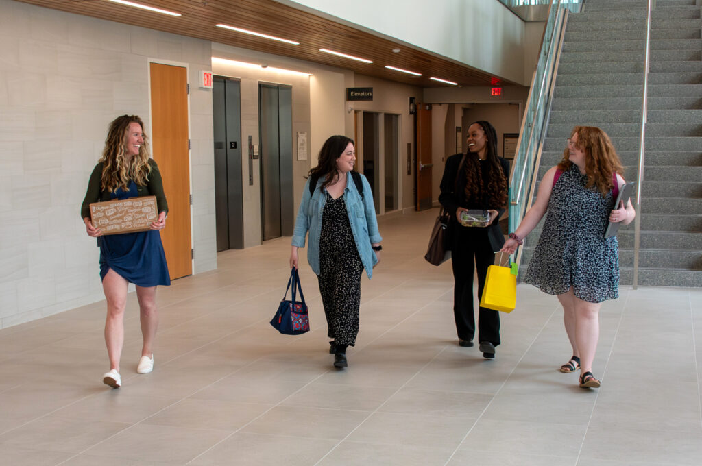 Four women carrying bags and boxes walk down a hallway together.