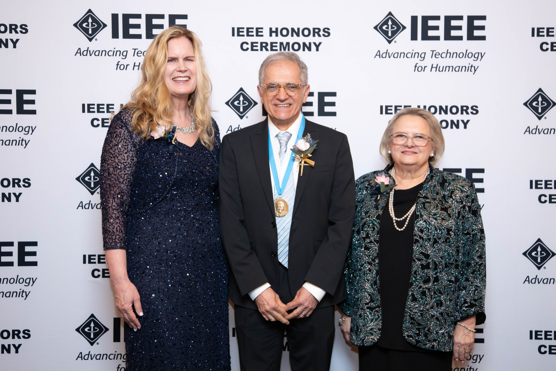 Kamal Sarabandi wears his medal and stands between two women in front of an IEEE background.