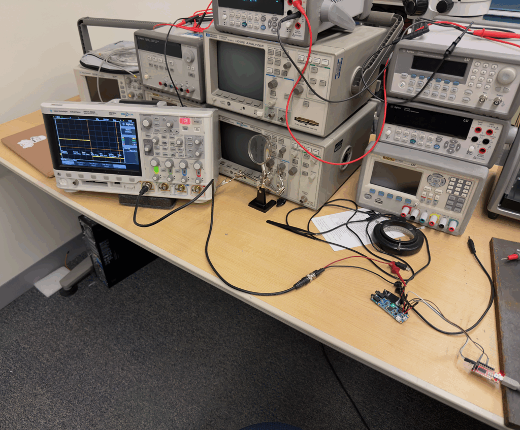 A workbench in a university lab with stacked electronic test equipment. Wires connect the devices to a small custom circuit board, suggesting active testing or debugging of an embedded system.