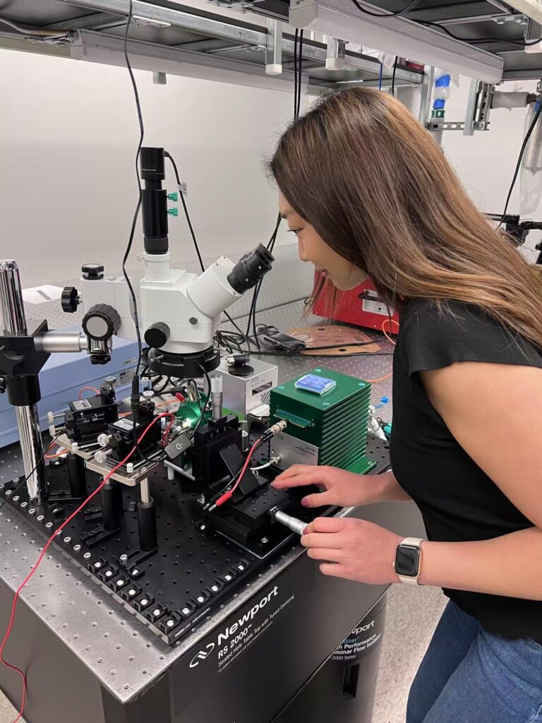 A woman adjusts some equipment while looking through a microscope.