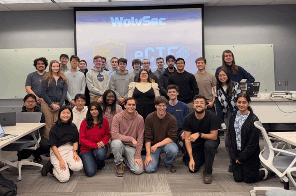 A group of about 30 students pose together in a classroom, smiling in front of a projector screen displaying the logos for WolvSec and eCTF.