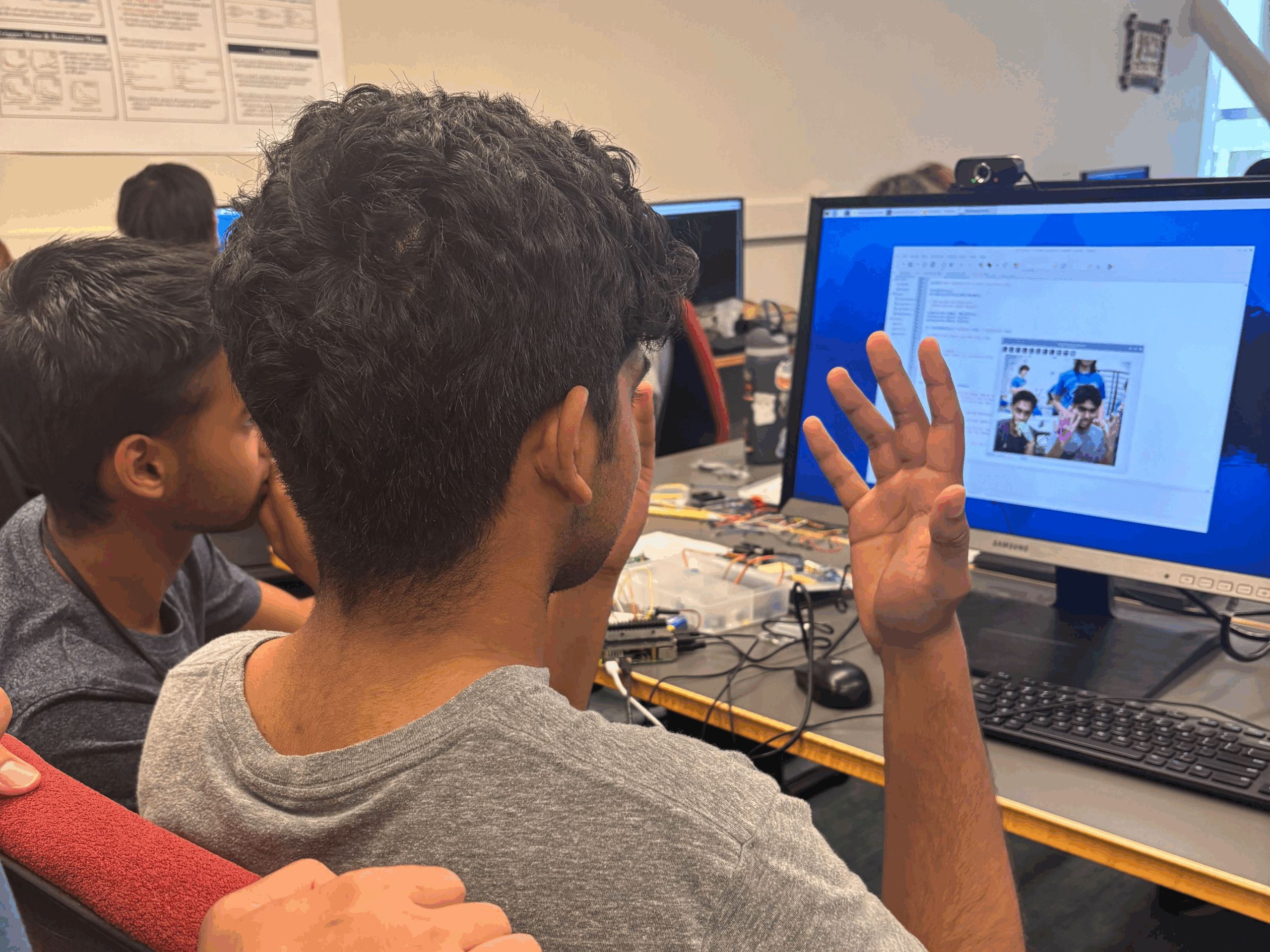 A high school student sits at a computer station in a lab, holding up one hand as an AI program on the screen detects and analyzes his gesture