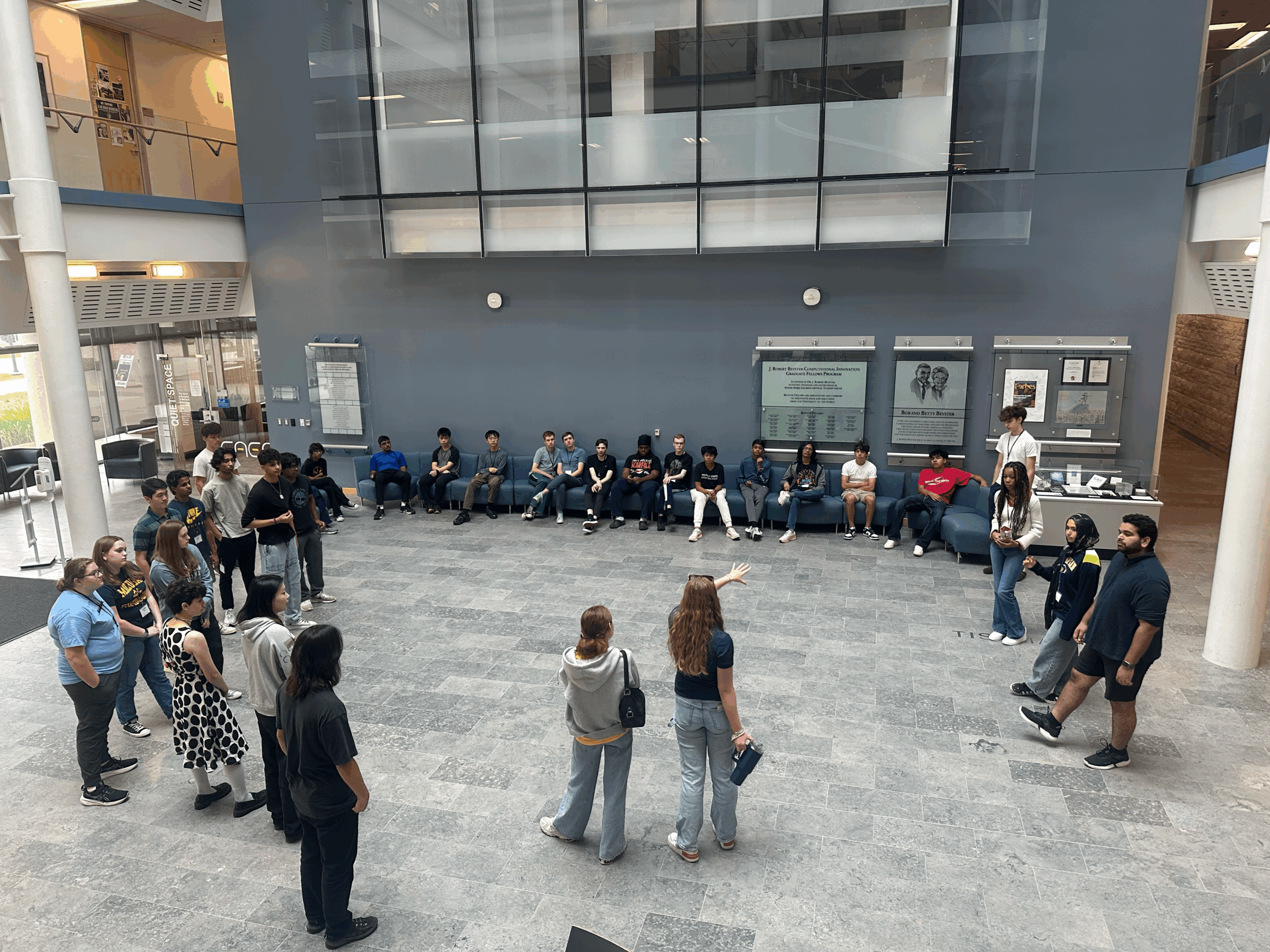 A large group of high school students gathers in a semicircle in a spacious campus atrium as a facilitator addresses the group.