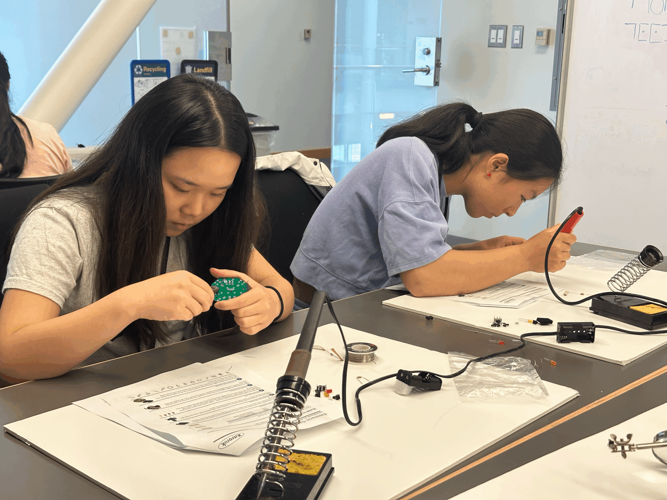 Two high school students are focused on assembling and soldering electronic circuit boards at a workshop table, surrounded by instruction sheets and equipment.