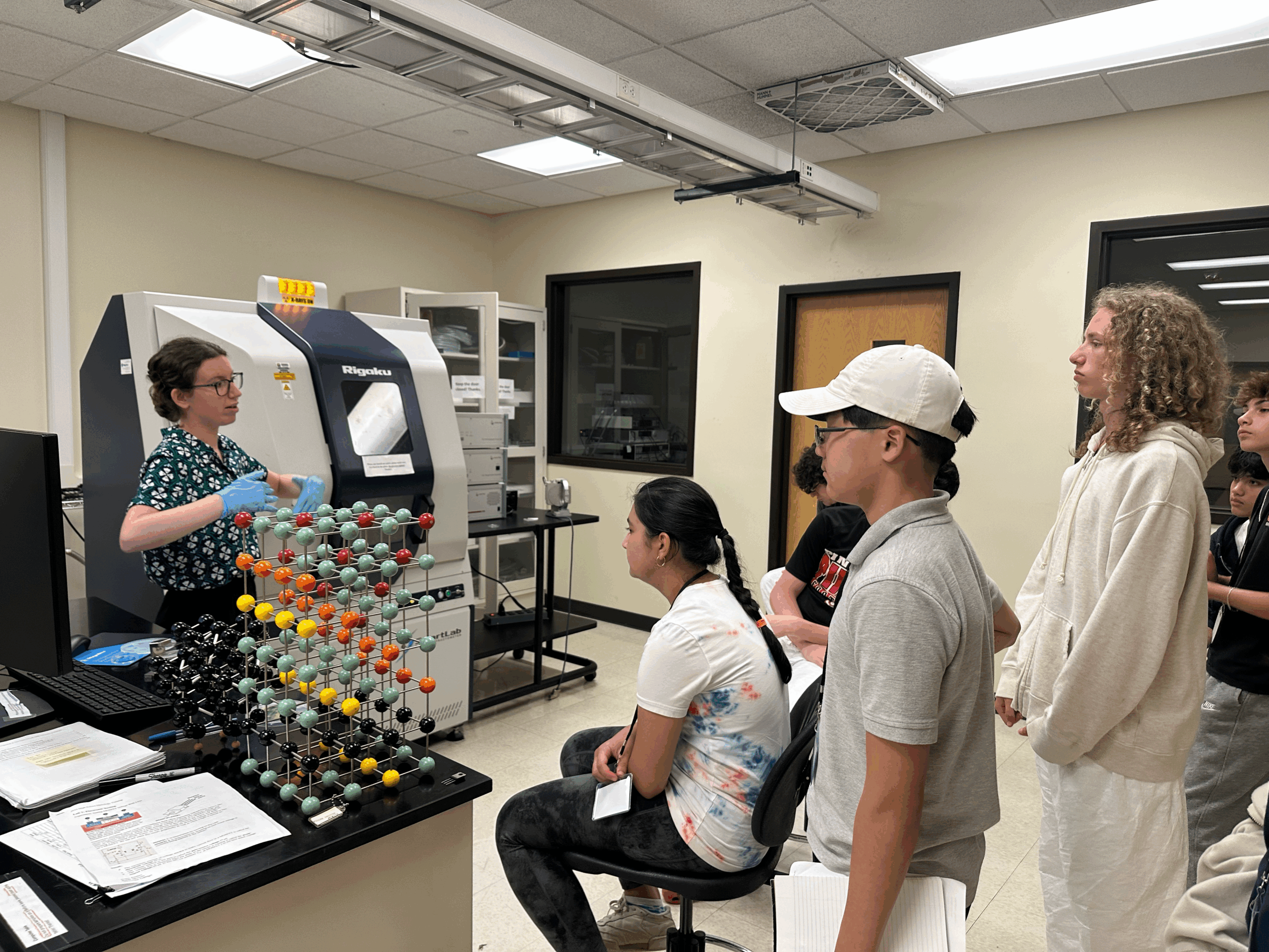 A researcher speaks to a group of high school students in a university laboratory with colorful molecular models and lab equipment nearby.
