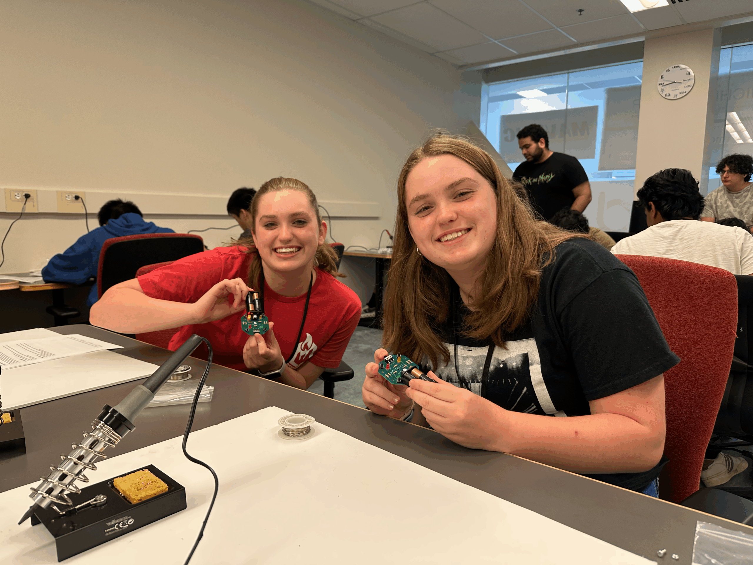 Two high school students sit at a workstation, proudly holding up small circuit boards they assembled, with soldering equipment and project materials visible on the table.