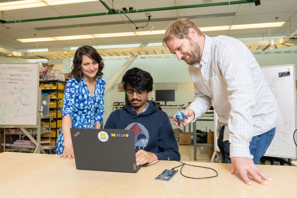 Three people stand around a laptop on a desk in a research lab. One of them holds an eyedrop tracking prototype.