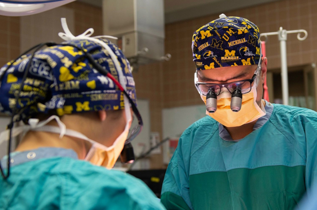 Two people in surgical garb are leaning forward, presumably performing surgery. One has magnifying glasses on. Both are wearing Michigan-branded caps.