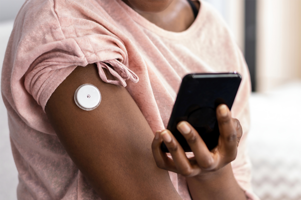 Person wearing a continuous glucose monitor on their upper arm and holding a smartphone.